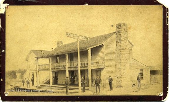 A yellow-toned picture of a large two-story building, with balcony decks and a stone chimney. Several men stand out front.