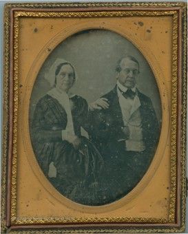 A black and white image of a middle-aged couple. The woman on the left wears a white cap, white shirt, and matching skirt and jacket. The man wears a waistcoat, bowtie, and jacket. The woman has a hand on the man's shoulder. They look into the camera