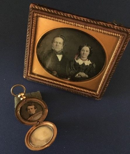 A framed image of a middle-aged man and woman, the man has graying hair and a jacket and bowtie, the woman has curled hair and a dress with a lacy white collar. To the left is a pocket watch-sized device with a picture of a young man inside