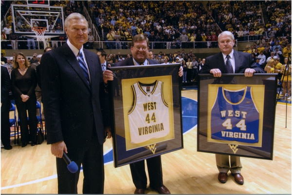 An elderly Jerry West standing on a court, next to him two men stand holding his number 44 west virginia jerseys in frames