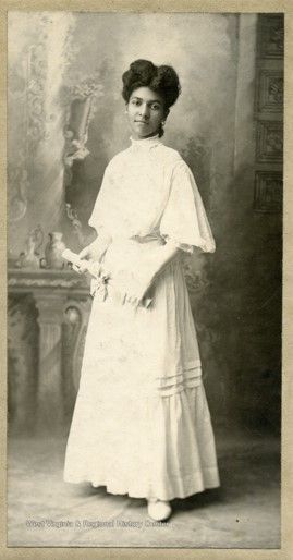 A woman with tan skin and dark hair wears a white high-necked gown and looks thoughtfully into the camera. In her hands, she holds a paper rolled up, with ribbon tied around the center