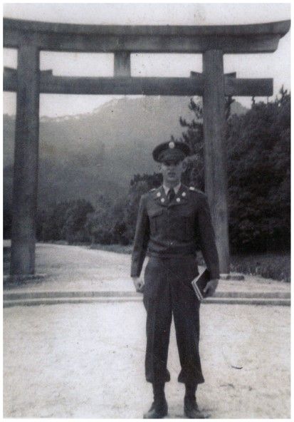 A young man stands in front of a rectangular archway. He is dressed in a military uniform, with a hat and boots. He is holding a small book in his left hand.