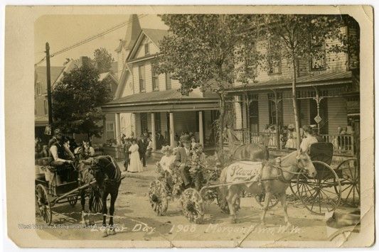 A road lined with two story houses, with people gathered on the porches and sidewalk. They watch a parade of wheeled floats pulled by horses.