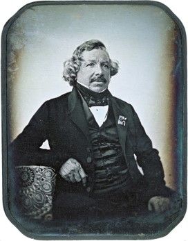 A black and white image of a man wearing a waistcoat, high-collared shirt, jacket, and a bowtie. He has parted, curly hair, a mustache and a neutral expression
