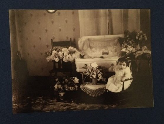 A sepia toned portrait of a small coffin surrounded by flowers and a toddler sat up next to it.