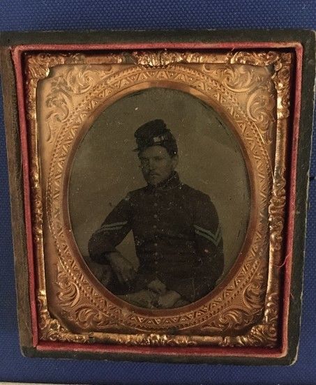 At tintype of a soldier wearing a uniform. The portrait is oval-shaped and the frame is an ornate bronzy-gold color. 