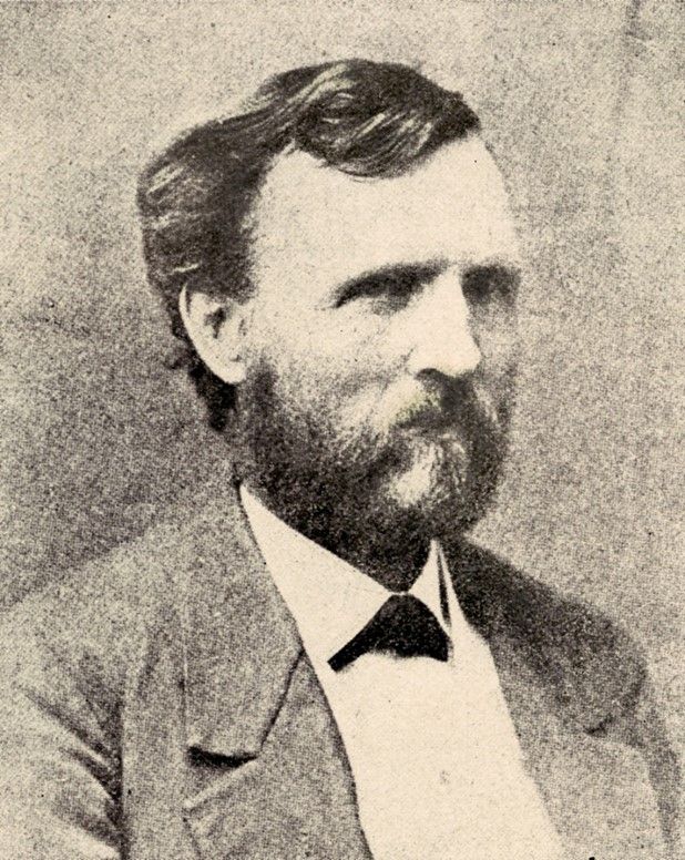 A middle-aged white man with a wave of dark hair and a short but full beard. His eyes are narrow and framed by low eyebrows. He is wearing a jacket and a white shirt 