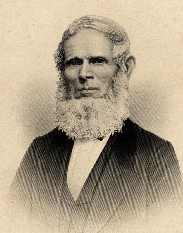 An older man with styled white hair and a long, full white beard. He has a slim face, high forehead, and pronounced cheekbones. He is wearing a vest, a jacket and a white shirt