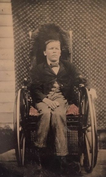 A white boy sits in a wheelchair, a hand resting on his lap. He wears slacks, a white shirt, a suit jacket and a bowtie, and looks thoughtfully into the camera. 