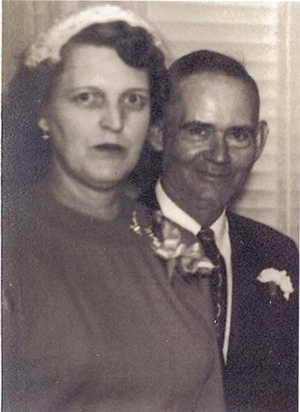 An image of a middle-aged woman and man, standing close to each other. The woman is wearing a dress, a white fascinator and has a corsage pinned over her chest. The man wears a suit, tie, and carnation pinned to his jacket. He's smiling, she looks serious