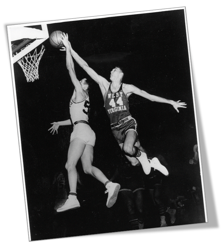 A photo of Jerry West jumping up to block a layup from the other team. They are both mid-air, with their arms outstretched toward the ball as it approaches the hoop