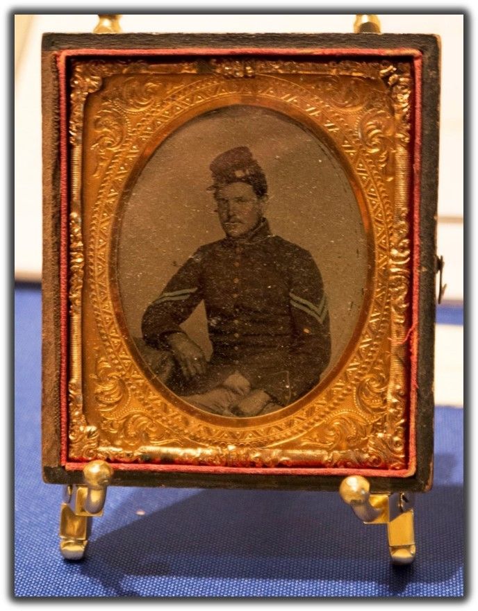 a soldier sits in uniform, looking into the camera. the frame is a gilded golden brown and warm red