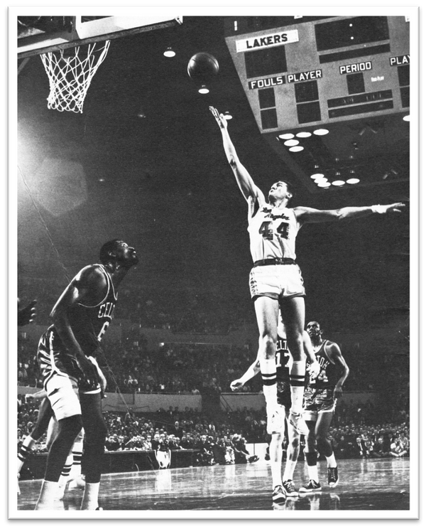 Jerry West mid-air on the court, jumping with both hands outstretched; one reaching up and one reaching out. two players from the opposing team surround him