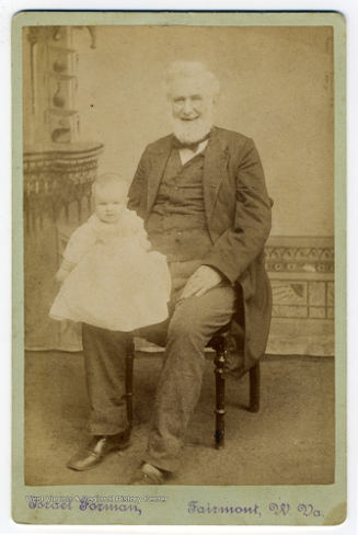 A cabinet card of an older man with white hair and a white beard, sitting in a wooden chair with a baby wearing a white gown on his lap.