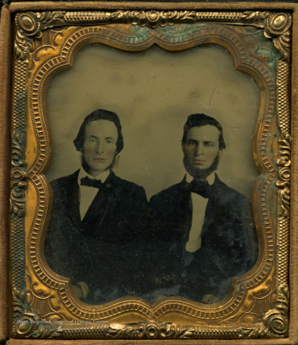 Two similar looking white men with dark hair, the one on the left has slightly longer hair and sideburns and the one on the right has a short beard. They both wear suit jackets, white shirts, and bowties.