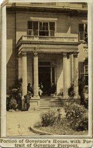 A yellowed image card of the front of the governor's house, a large building with four tall columns and steps leading to a large door. A stone or brick pathway leads through a garden and to the base of the steps.