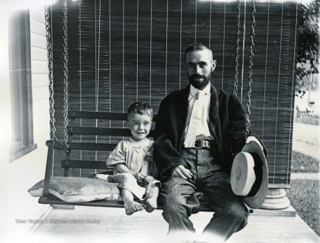 A man with a shaved head wearing a jacket holds a brimmed hat and sits on a wooden porch swing. Next to him, a toddler sits barefoot. they both smile at the camera 