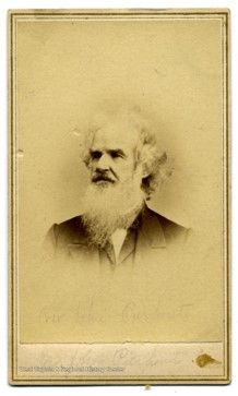 A yellowed image of an older man, with a long white beard, and medium length grayish white hair on the sides of his head. He wears a jacket and stares to his right. 