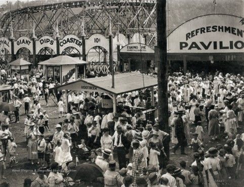 A large crowd of hundreds of people stand around a roller coaster. 