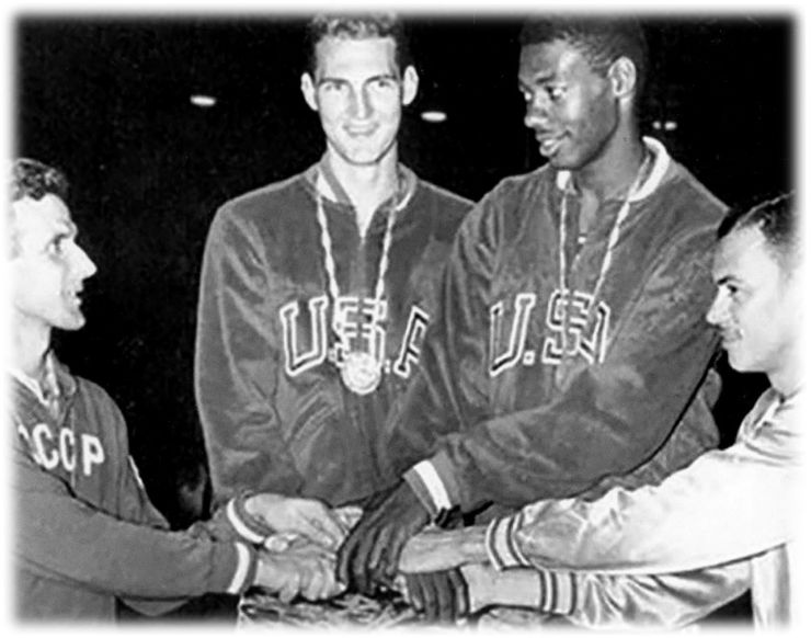 Four men stand and put their hands in the center of the huddle. in the middle are two men wearing USA sweatshirts and medals. They are smiling. The one on the left is Jerry Lewis, and the one on the right is Oscar Robertson
