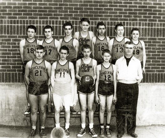 A black and white phot of a middle school basketball team, wearing their uniforms and standing in front of brick wall. Jerry West wears a number 12