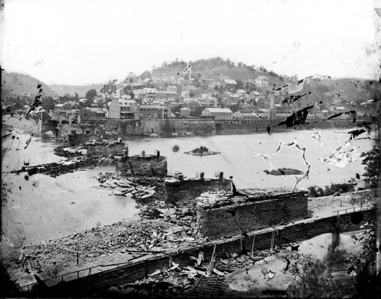 Black and white image of a bridge destroyed by Confederate troops. Pieces of splintered wood sit in piles in the water below