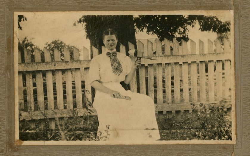 A young woman with her hair pulled back in a white dress and a plaid necktie holds a stereoscope in her left hand. She sits in front of a worn picket fence. Behind the fence, are two trees.