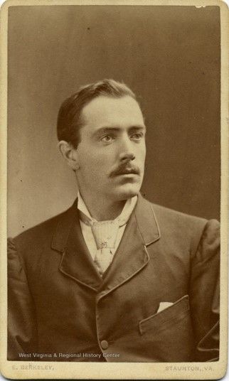 A young adult man, with neatly styled dark hair, a mustache, and high cheekbones wears a high-collared white shirt, necktie and jacket. He looks off to his left 