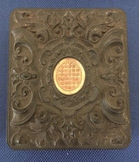 A tall rectangular, dark brown photo case with a swirling inset design, and a gold oval in the middle 