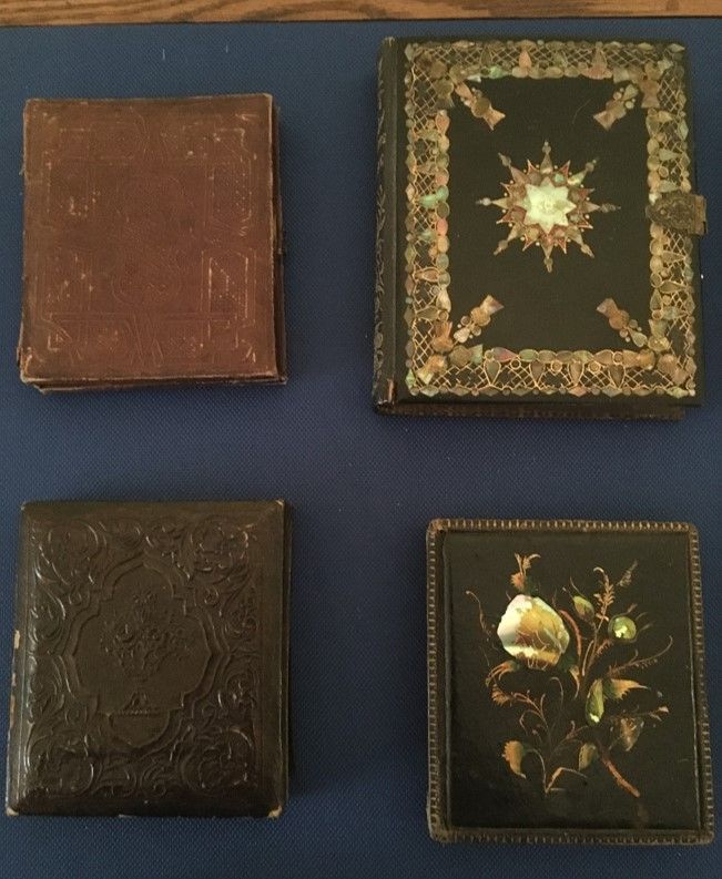 Four photo cases sit against a blue background. The upper left is a worn light brown, the bottom left is a dark brown, the top right is a dark color with gold ornamentation around the border and a sunburst, bottom right has a detailed floral design 