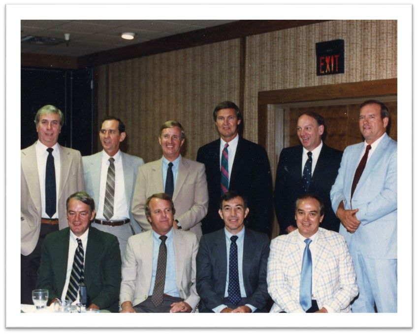 A group of ten middle-aged men wearing suits and ties stand in two rows. They smile at the camera 