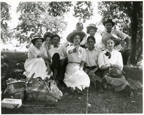A black and white image of several people, mostly women, sitting in the grass with picnic baskets, and wearing light colored clothes. The woman in the middle points at the camera
