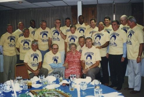 A group of several men pose with Ann, all wearing pale yellow shirts with her face on them. It is ann's 90th birthday and they are all smiling 