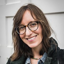 A white woman with glasses and shoulder length brown hair smiles in front of a white door
