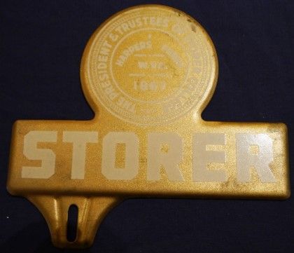 A gold Storer College license plate tag. The top is a circle with a crest and the bottom is a rectangle that reads "STORER."
