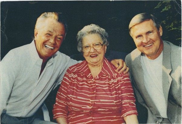 Two middle-aged men pose with an elderly woman. The man on the left wears a button down and has his arm around her. The man on the left wears a jacket. They are all smiling