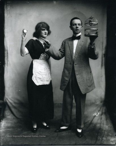 A woman and man stand in front of a cloth backdrop: the woman is wearing a dark dress with short sleeves and a white apron, and is holding a stopwatch up; the man is holding a small birdcage in one hand and pointing to his right with the other. 