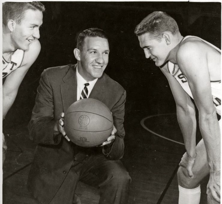 Black and white, two players stand on either side of a coach. The players weird uniforms and crouch down smiling to look at the ball the coach holds in his hands. He is smiling and on one knee.