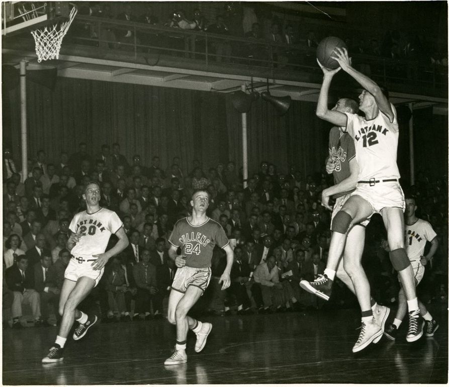 Mid-game, Jerry West is mid-air in a jump shot. Other players watch on. One lunges for the ball
