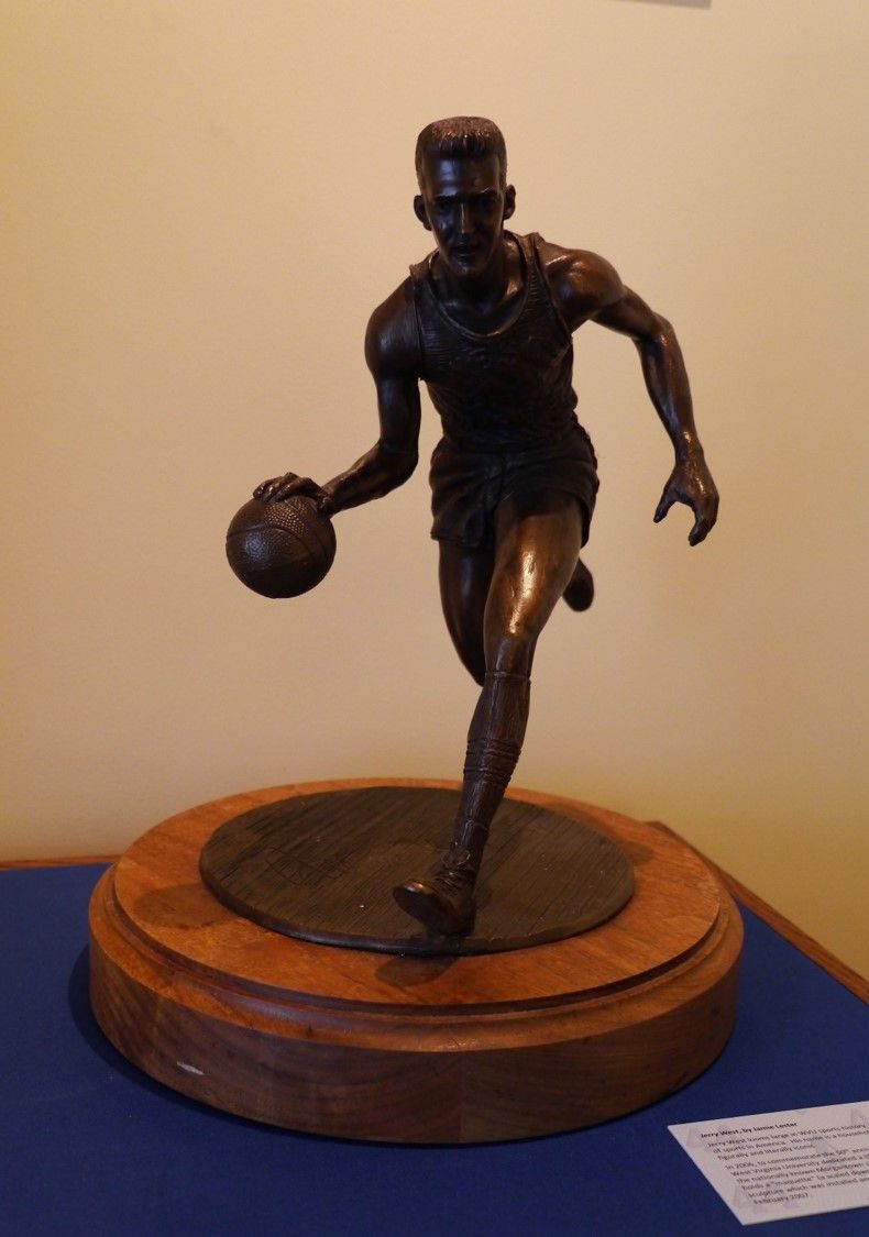 A small bronze statue on a wooden base. It depicts Jerry West dribbling a ball, mid-motion. 