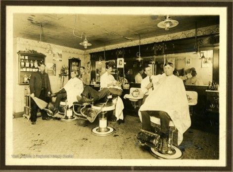 Three men sit barber chairs in front of a mirror. They wear white capes over their clothes. 