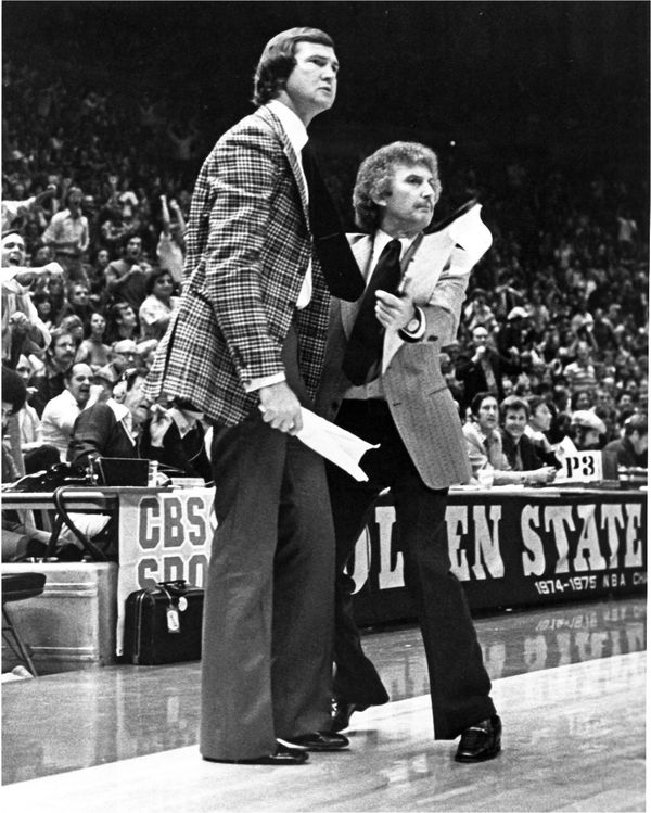 Jerry west, with longer hair and wearing a plaid suit jacket and dress pants, stands to the left of a shorter man with a jacket, tie, goatee and medium length gray hair.