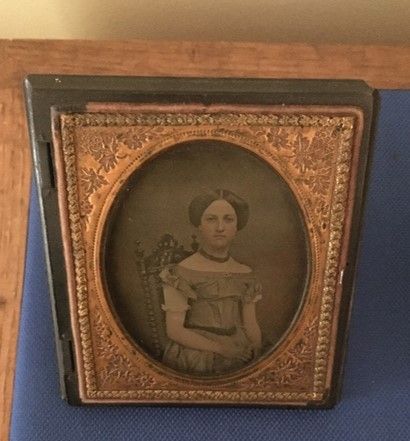 An oval shaped ornately framed photo of a young woman, wearing her hair parted in the center and in an updo, and a wide-necked dress