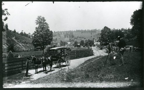 An image of a dirt road next to a fence. Trees and hills are in the distance, and a car pulled by horses stands in the foreground. 