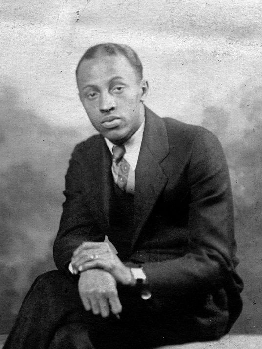 A black man wearing a suit sits with a leg crossed, and his hands on his lap. He looks thoughtfully into the camera 
