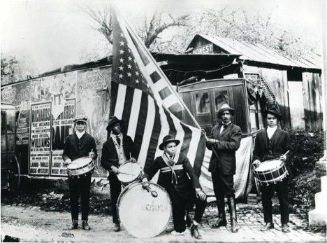 A group of young black men wearing suits and hats hold various drums. One man in the center holds up a large american flag