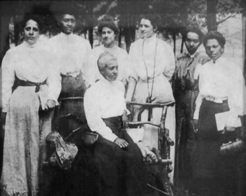 A group of women in white shirts and full skirts stand outside. One sits in front, looking away from the camera