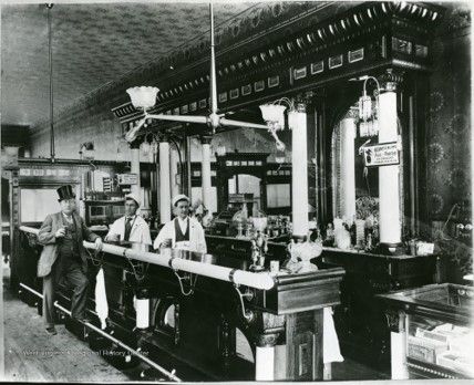 An ornate bar, with two bartenders behind wearing white coats and hats, a man with a top hat stands with one foot up against the bar.