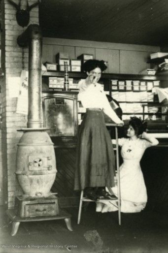 Two women stand in a store next to a wood burning furnace/oven. The woman on the left is standing on a metal stepladder and pointing at the camera, and the woman on the right sits on the floor with a hand behind her head and gazes up at the other woman