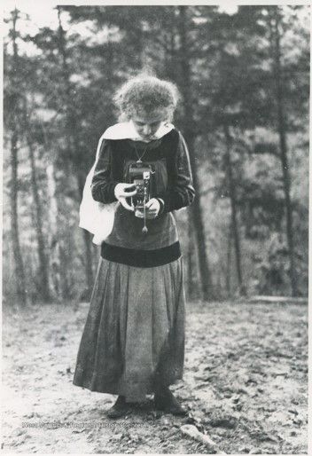 A young woman in an ankle-length dress and curly hair in an updo stands outside. In her hands she holds a brownie bellows camera and looks down at it